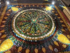 A close-up, overhead view of a large, intricate circular mosaic or mandala made entirely of colorful polished stones, crystals, and glass pieces.