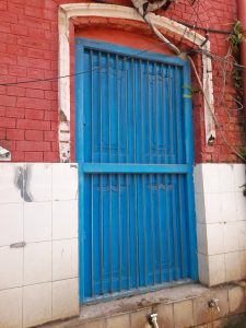 A vibrant blue metal door with horizontal bars is set against a red brick wall. 