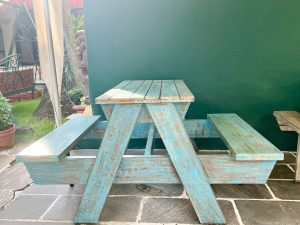 A weathered, peeling blue wooden picnic table with benches sits on a dark stone patio against a solid teal-green wall.
