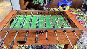 A wooden foosball table with red and blue players stands ready for a game on a brightly colored, abstract-patterned carpet.