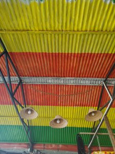 
A vibrant corrugated metal roof with horizontal stripes of yellow, green, red, and orange, featuring three straw hats with light bulbs hanging from a black metal frame.