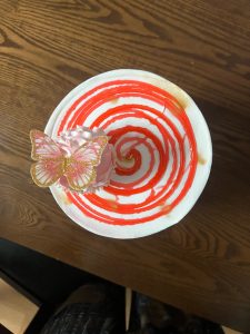 A close-up, overhead shot of a birthday cake,  decorated with a light pink frosting swirl and a delicate, shimmering butterfly ornament. The cake sits on a white plate with a striking red and gold spiral pattern.
