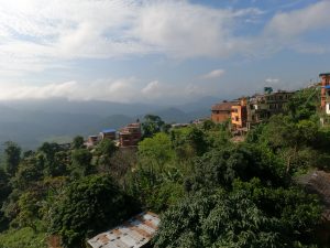 A scenic hillside landscape featuring a cluster of colorful houses among lush green trees and vegetation. The background shows rolling hills and distant mountains under a partly cloudy blue sky.
