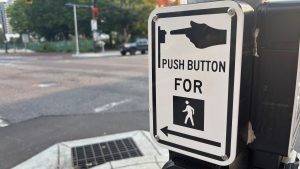 On the right, a sign on a post that shows a hand pushing a button, and it says "Push button for" and below that is a silhouette of a human walking, and an arrow. The background is a blurred street.