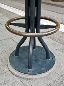 A dark metal bar stool base with a circular footrest, anchored to a concrete base on a tiled ground, likely outdoors near tracks. Captured from Providence Park, Portland.