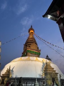 A large stupa with a distinctive golden top, adorned with colorful prayer flags, is illuminated against a twilight sky. 