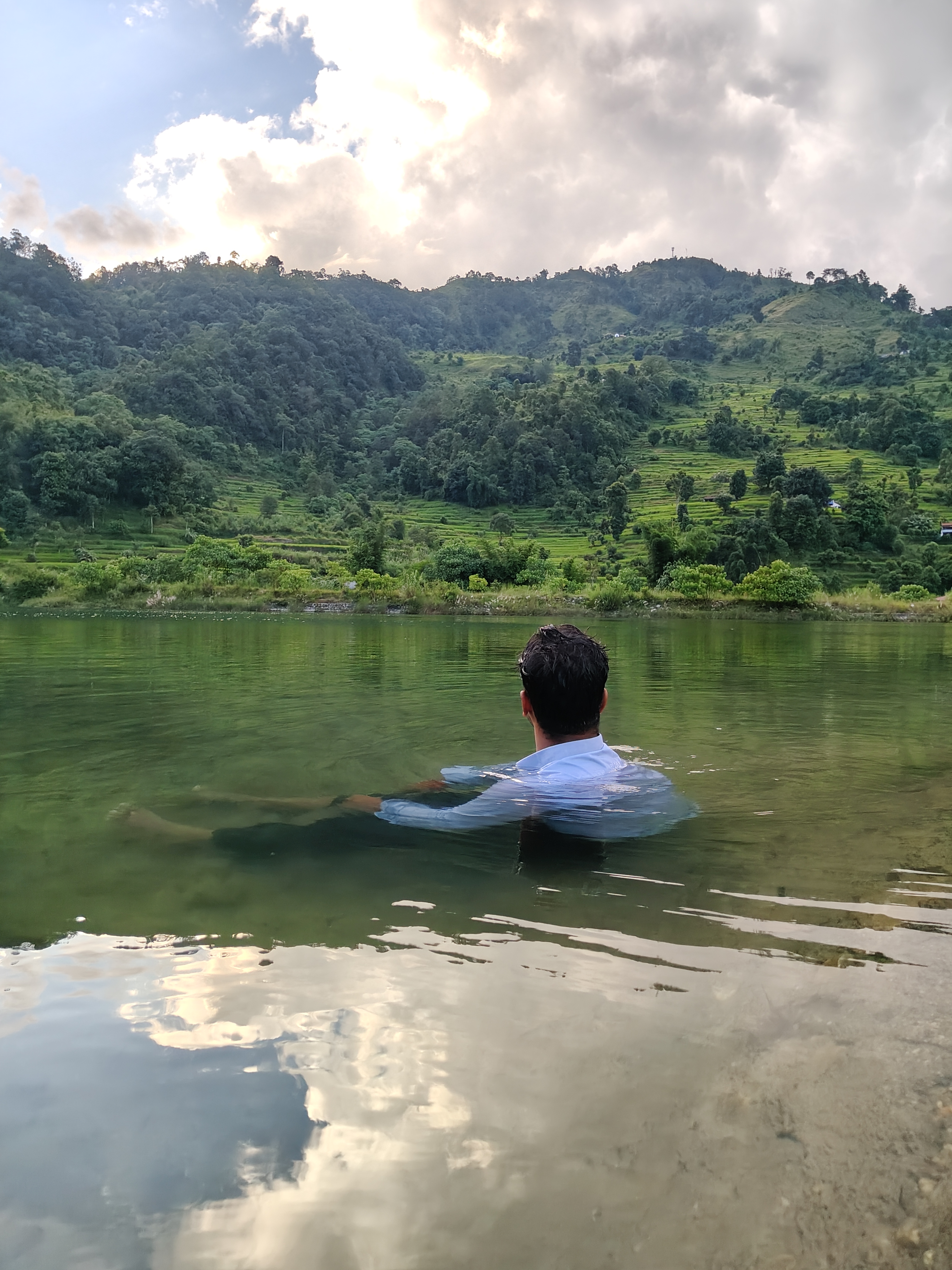 A person is sitting in shallow water, partially submerged, with their back facing the camera. 