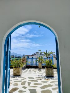 View through a blue arched doorway onto a rooftop terrace in Da Nang, Vietnam. The terrace has potted plants, patterned stone flooring, and a blue fence, with city buildings and mountains visible under a bright blue sky.