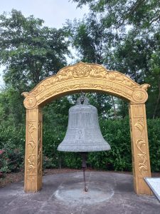 A large, ornate bell made of a mottled gray material is suspended from a decorative archway that is painted in a bright gold color. 