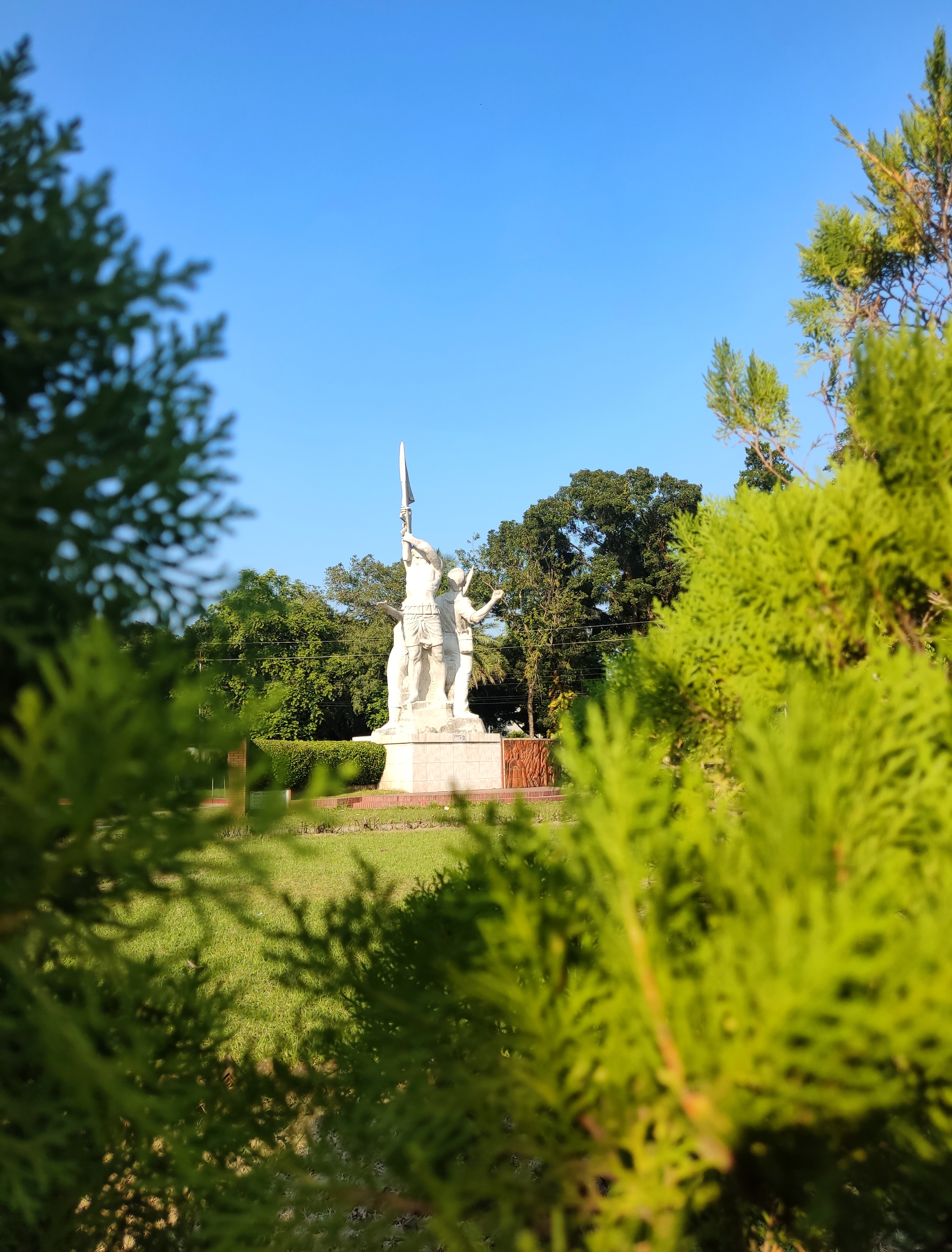 A historical Shaheed Minar monument standing tall in a lush green garden, framed by trees and bright blue sky. The white stone sculpture symbolizes courage, freedom, and sacrifice, making it a cultural landmark and patriotic heritage site. Perfect for travel, history, and educational content.