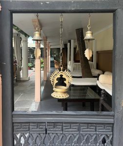 A carved wooden doorway frames a patio with white seating, brass Dalucha lamps, and hanging bells.