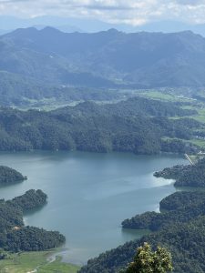 A panoramic view of a serene lake surrounded by lush green mountains. The water is calm and reflects the blue sky and fluffy white clouds above. Various shades of green from the trees and hills create a picturesque landscape, with small patches of farmland visible in the valley below. The scene conveys a sense of tranquility and natural beauty.
