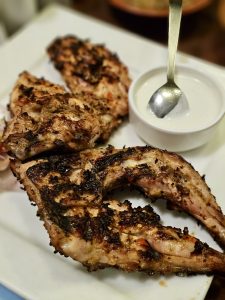 Grilled chicken pieces with charred edges arranged on a white plate, served with a bowl of creamy white dipping sauce and a metal spoon. 