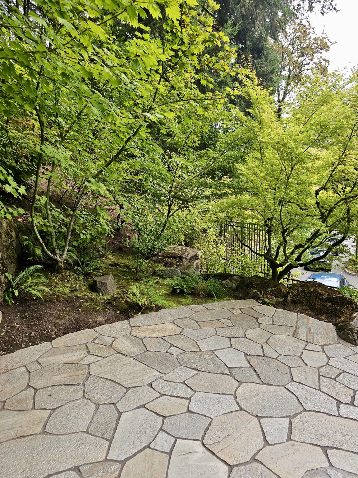 A stone-paved area surrounded by small green trees and mossy rocks at the Portland Japanese Garden. A blue car is visible in the background beyond the garden fence.