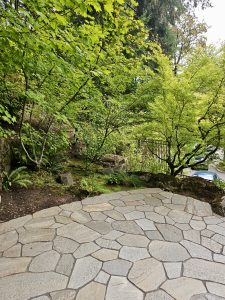 A stone-paved area surrounded by small green trees and mossy rocks at the Portland Japanese Garden. A blue car is visible in the background beyond the garden fence.