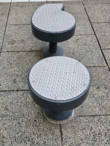 Two dark gray, metal-topped stools or seating elements with a textured, diamond plate pattern on their surfaces, set on a concrete ground. Captured from Providence Park, Portland.