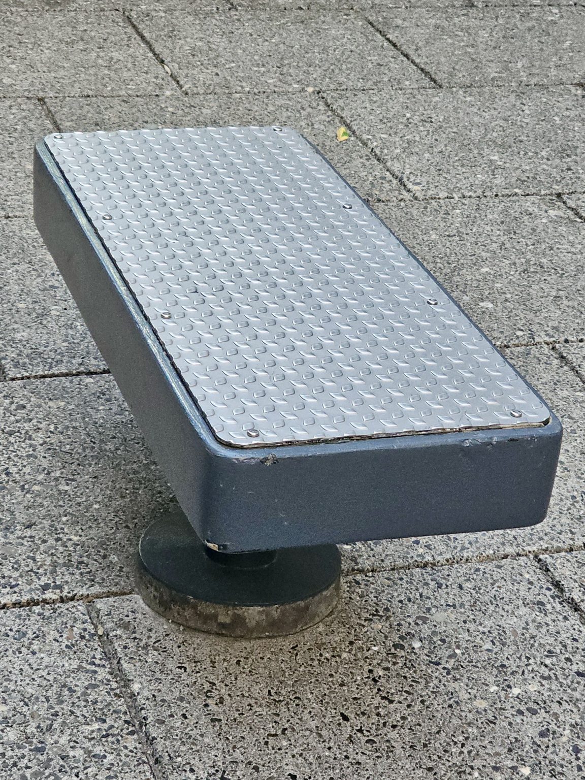A metal-topped, dark gray outdoor bench with a textured, diamond-plate surface, mounted on a round base on a paved area. Captured from Providence Park, Portland.