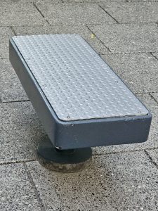 A metal-topped, dark gray outdoor bench with a textured, diamond-plate surface, mounted on a round base on a paved area. Captured from Providence Park, Portland.