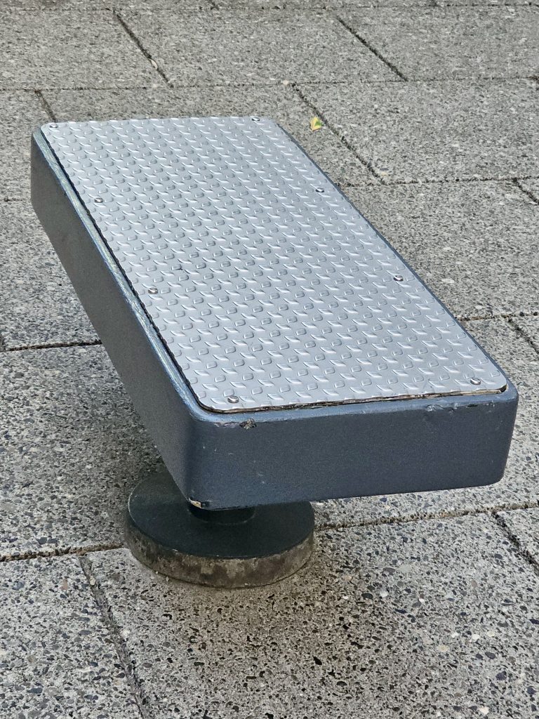 A metal-topped, dark gray outdoor bench with a textured, diamond-plate surface, mounted on a round base on a paved area. Captured from Providence Park, Portland.