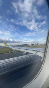 A view from an airplane window showing the wing and an airport runway during what looks like a takeoff or landing. The sky is bright blue with white clouds