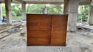A wooden storage cabinet with a textured surface featuring geometric designs, positioned outdoors on a dirt ground littered with debris.