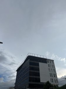 A low-angle shot of a modern, multi-story office building with a dark glass facade and a white section. The sky above is cloudy.