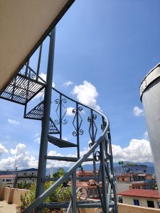 A spiral staircase made of metal curves upward against a bright blue sky with fluffy white clouds