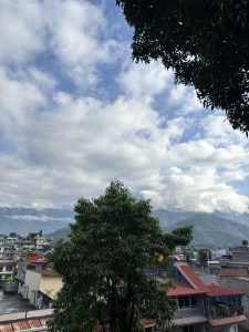A view from a high vantage point, capturing a cityscape with buildings featuring colorful rooftops, green vegetation, and distant mountains under a partly cloudy sky.