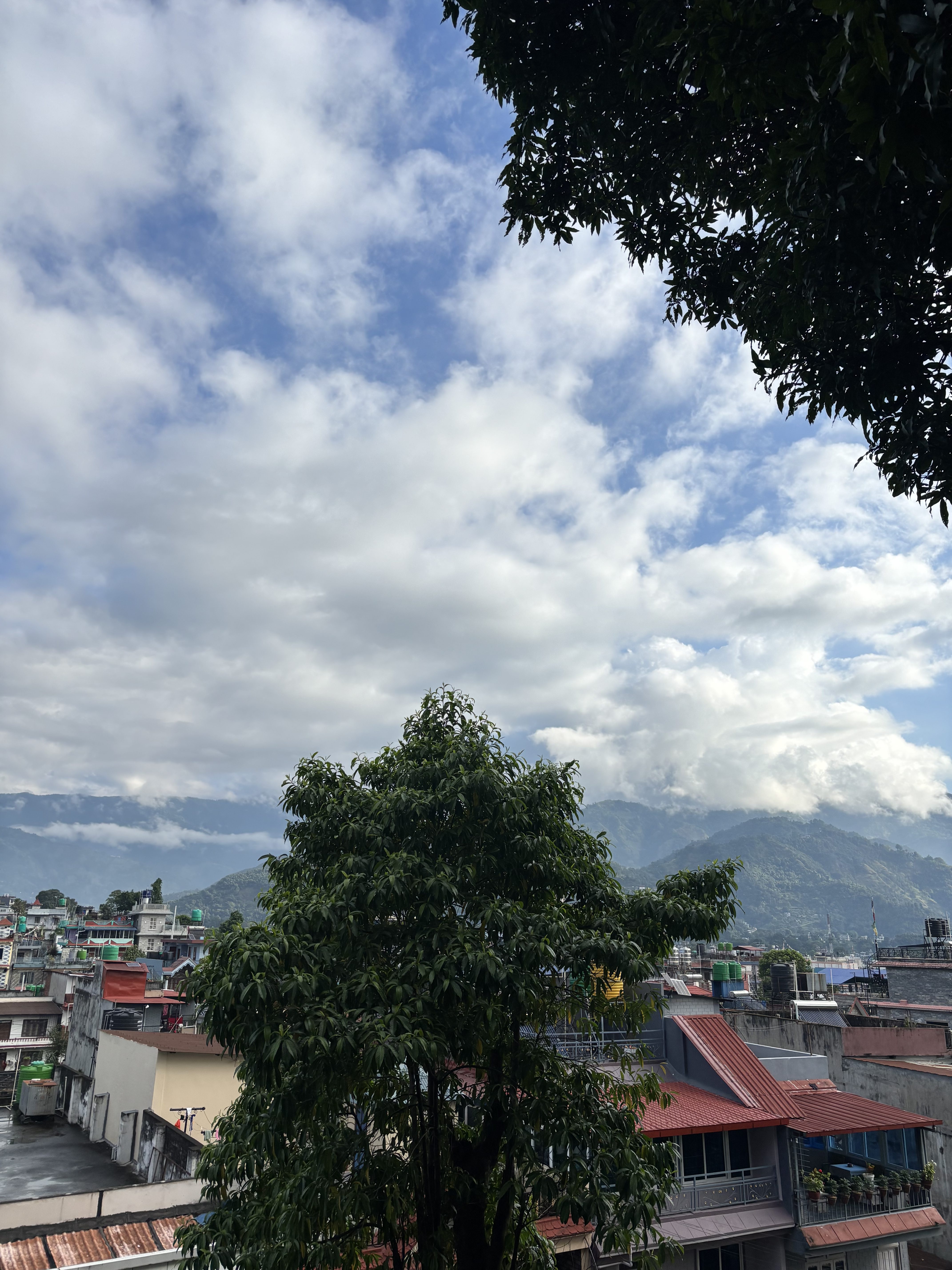 A view from a high vantage point, capturing a cityscape with buildings featuring colorful rooftops, green vegetation, and distant mountains under a partly cloudy sky.