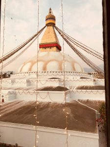 A large, white stupa with a gilded spire and colorful prayer flags is shown from an elevated viewpoint, with string lights in the foreground.