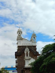 Rajwada of Ichalkaranji. A decorative building facade with intricate architectural details, featuring arched openings and ornate pinnacles. The structure is predominantly white with some dark stone elements and is partially obscured by lush green foliage on the right. The sky above is partly cloudy, showcasing blue and white hues.