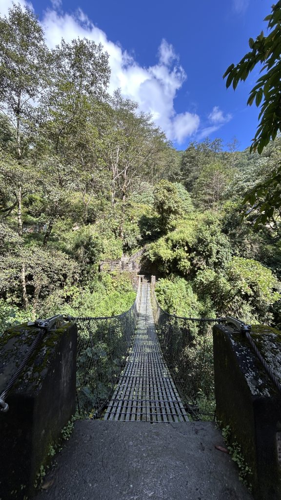 A suspension bridge made of wooden planks and supported by metal cables stretches across a lush, green valley.