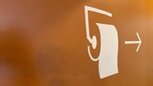 On an orange coloured wall, a stencil painting of a toilet paper roll, with an arrow pointing away the direction.