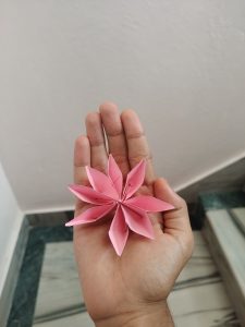 A hand holding a pink paper-made flower with multiple pointed petals, set against a light-colored wall and a hint of patterned flooring in the background.