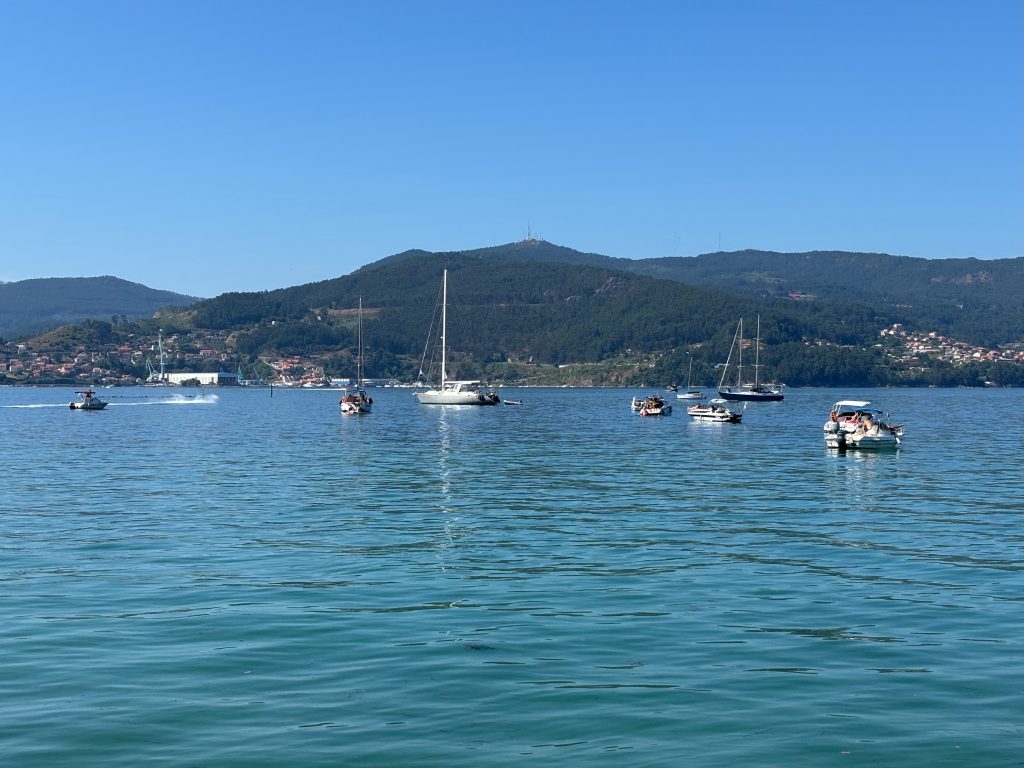 Small boats on a sunny day in the coast of Vigo, in the distance water side and hillside settlements and trees, the highest telecoms masks on the highest summit.