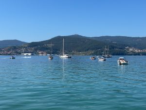 Small boats on a sunny day in the coast of Vigo, in the distance water side and hillside settlements and trees, the highest telecoms masks on the highest summit. 
