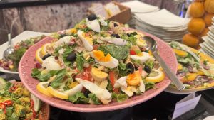 A large pink platter holds a mixed green salad that is generously topped with quartered hard-boiled eggs, olives, and lemon slices.