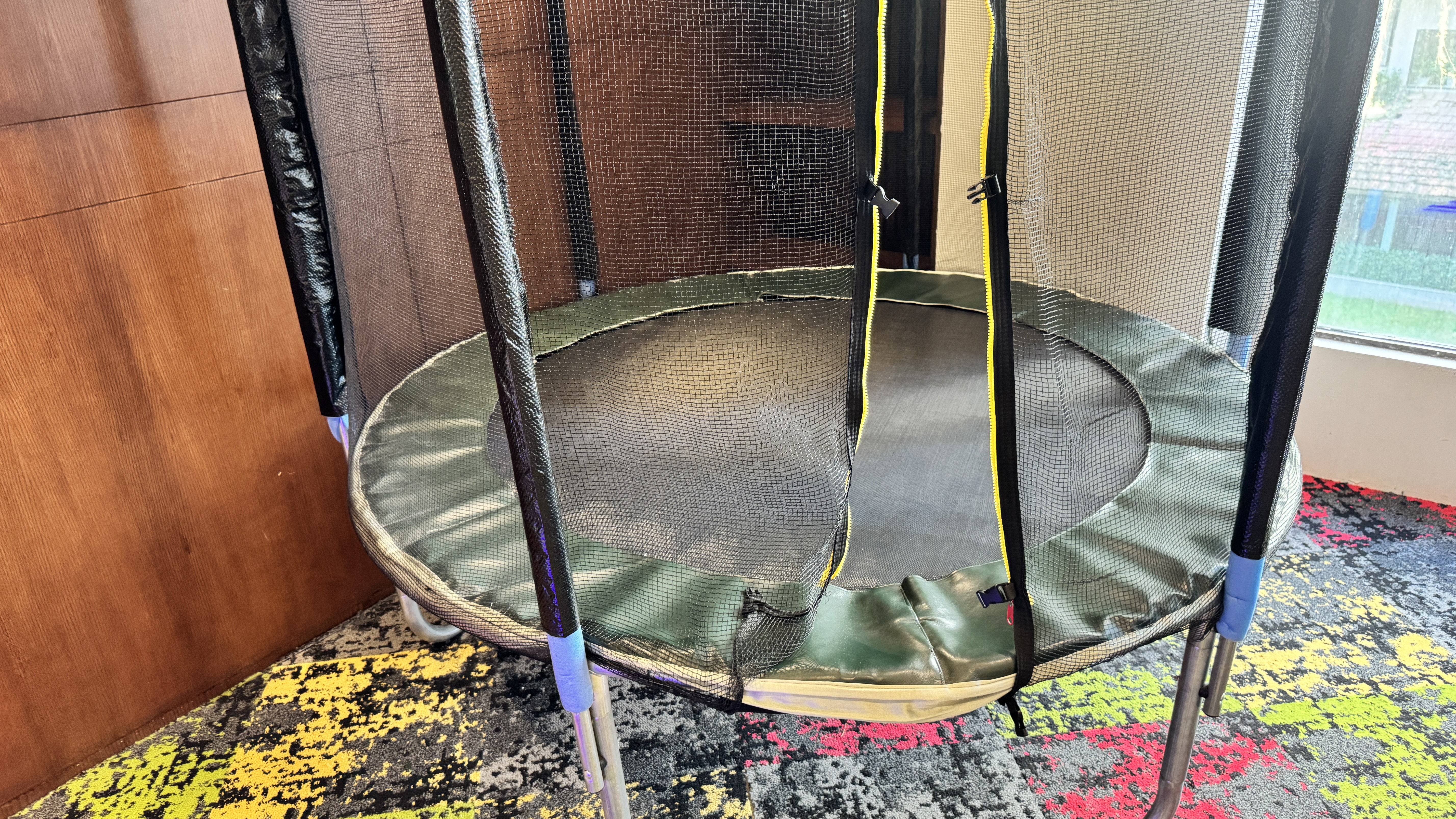 A small, indoor children's trampoline with a black safety net sits in the corner of a room with a colorful carpet and wood-paneled wall.