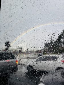 Rainy day view through a car window with raindrops, a faint rainbow in the cloudy sky, and cars in the foreground.