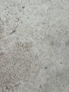 A close-up view of a textured gray concrete surface, featuring various specks of dirt and debris.