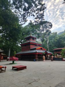 

A serene temple with red walls and a gold-accented roof, surrounded by greenery, featuring a pink lotus sculpture and a stone courtyard under a partly cloudy sky.