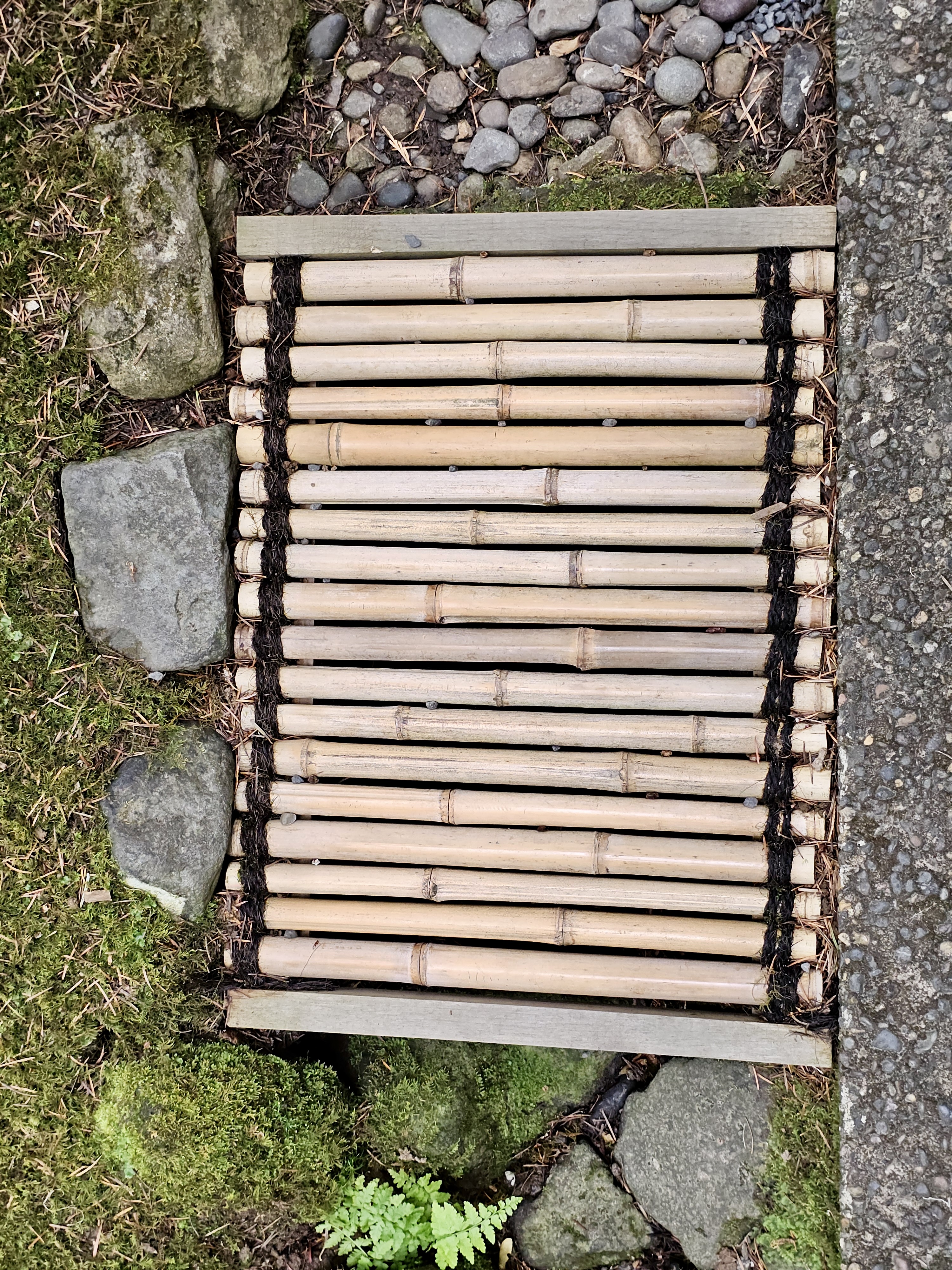 A close-up of a bamboo mat drain framed by moss and stones, used in traditional Japanese landscaping at the Portland Japanese Garden.