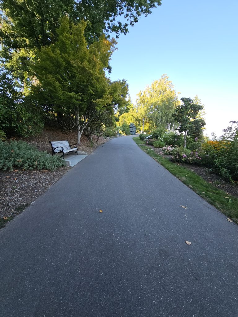 A peaceful pathway lined with trees on both sides, leading into the distance. A wooden bench is positioned along the path on the left, surrounded by lush greenery and colorful flower beds. The sky is clear with a hint of blue. Shot at Pittock Mansion, Portland.