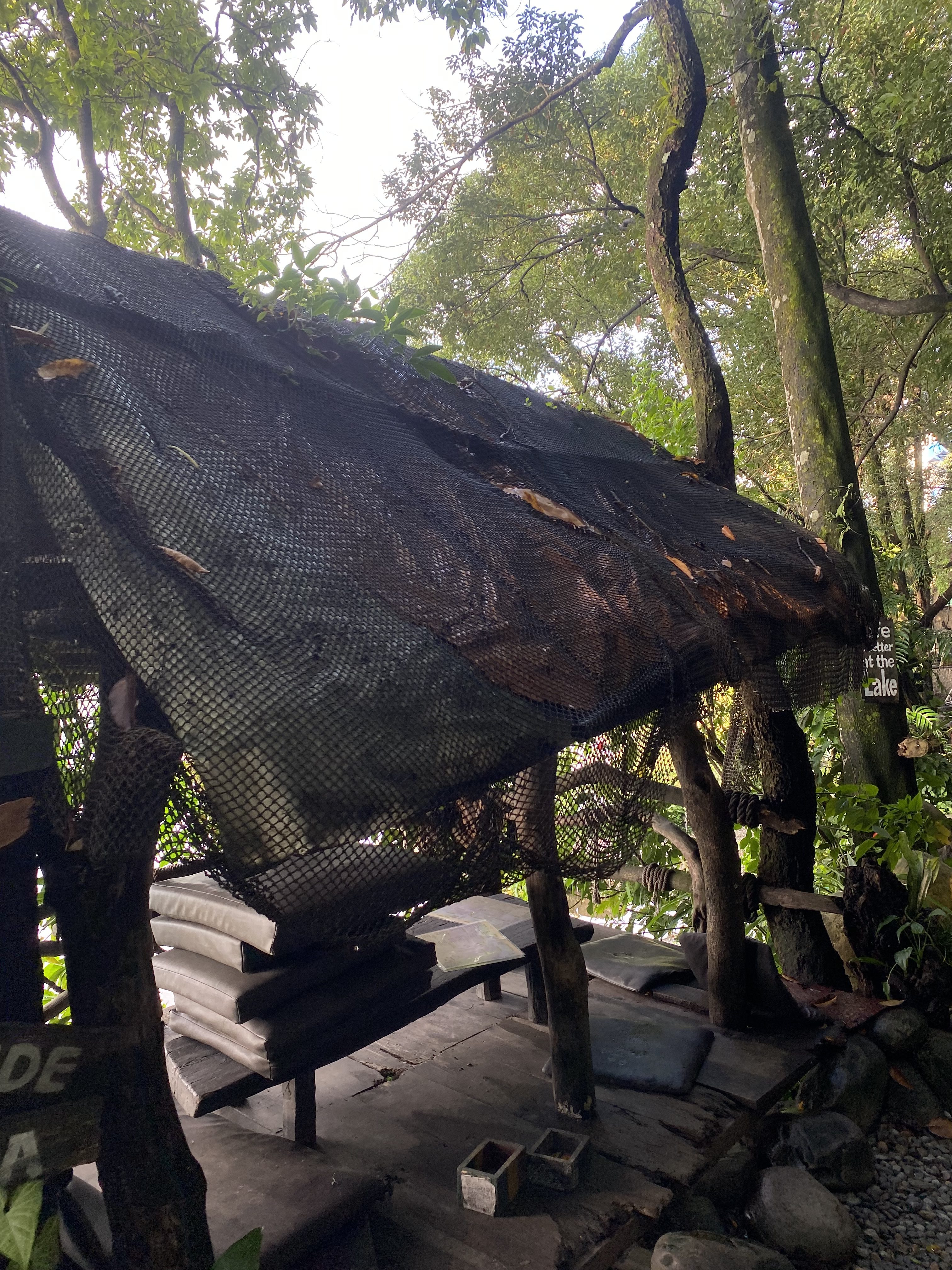 A rustic, open-air shelter made from wooden beams and a netted roof, surrounded by lush green foliage. 