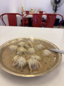 A bowl of dumplings in a light broth, garnished with green herbs and spices, is placed on a white table.