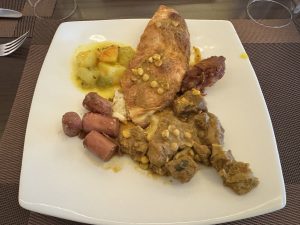 A white plate holds a varied breakfast of beef curry, sliced sausage, a vegetable stew, and a piece of folded flatbread.