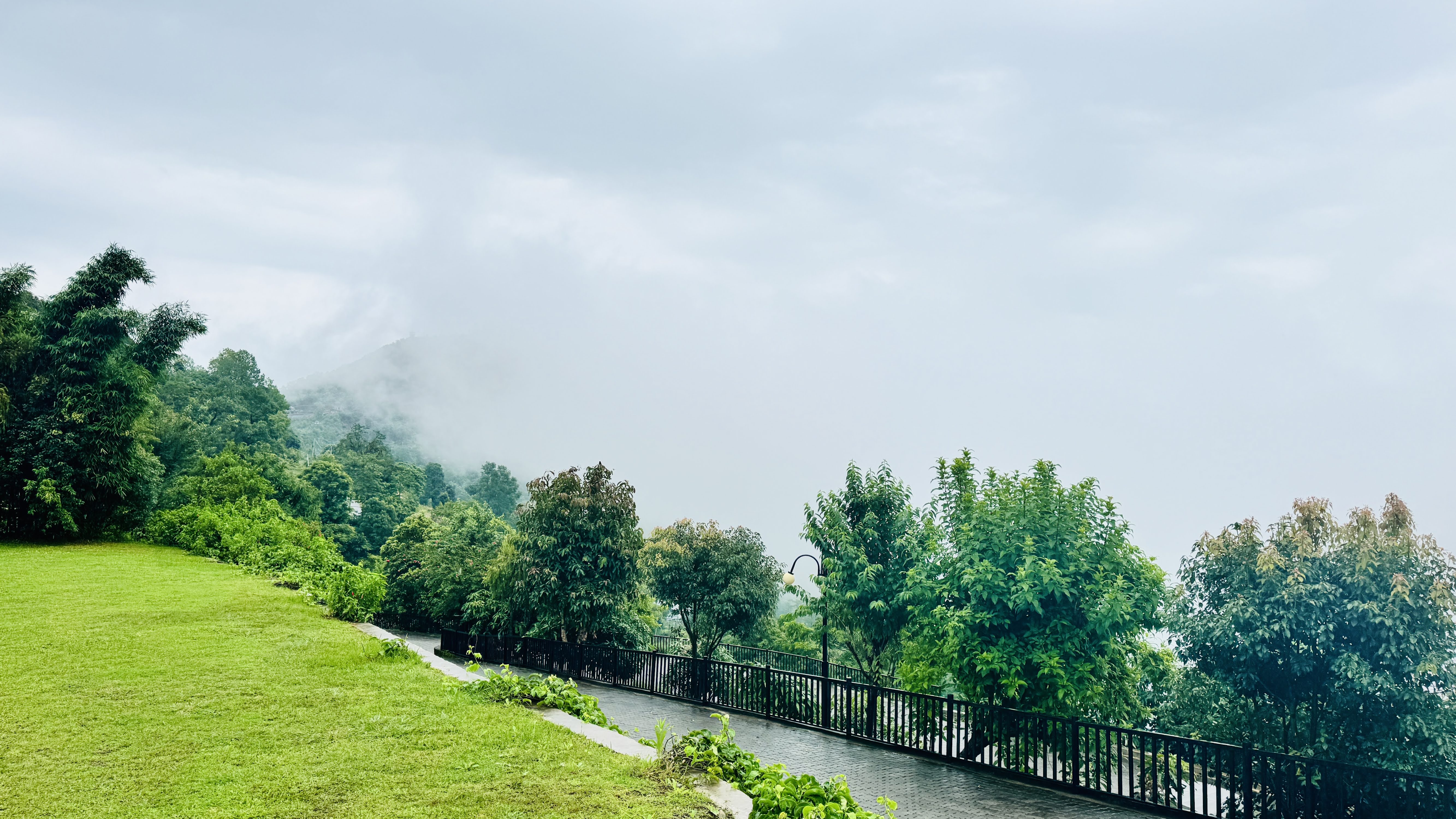 A scenic view featuring lush greenery and trees under a cloudy sky. In the foreground, a well-maintained lawn transitions into a pathway that is bordered by a black metal fence