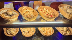 A bakery shelf displays a variety of fresh bread rolls, including some with seeds and glazes, all arranged in wicker baskets.
