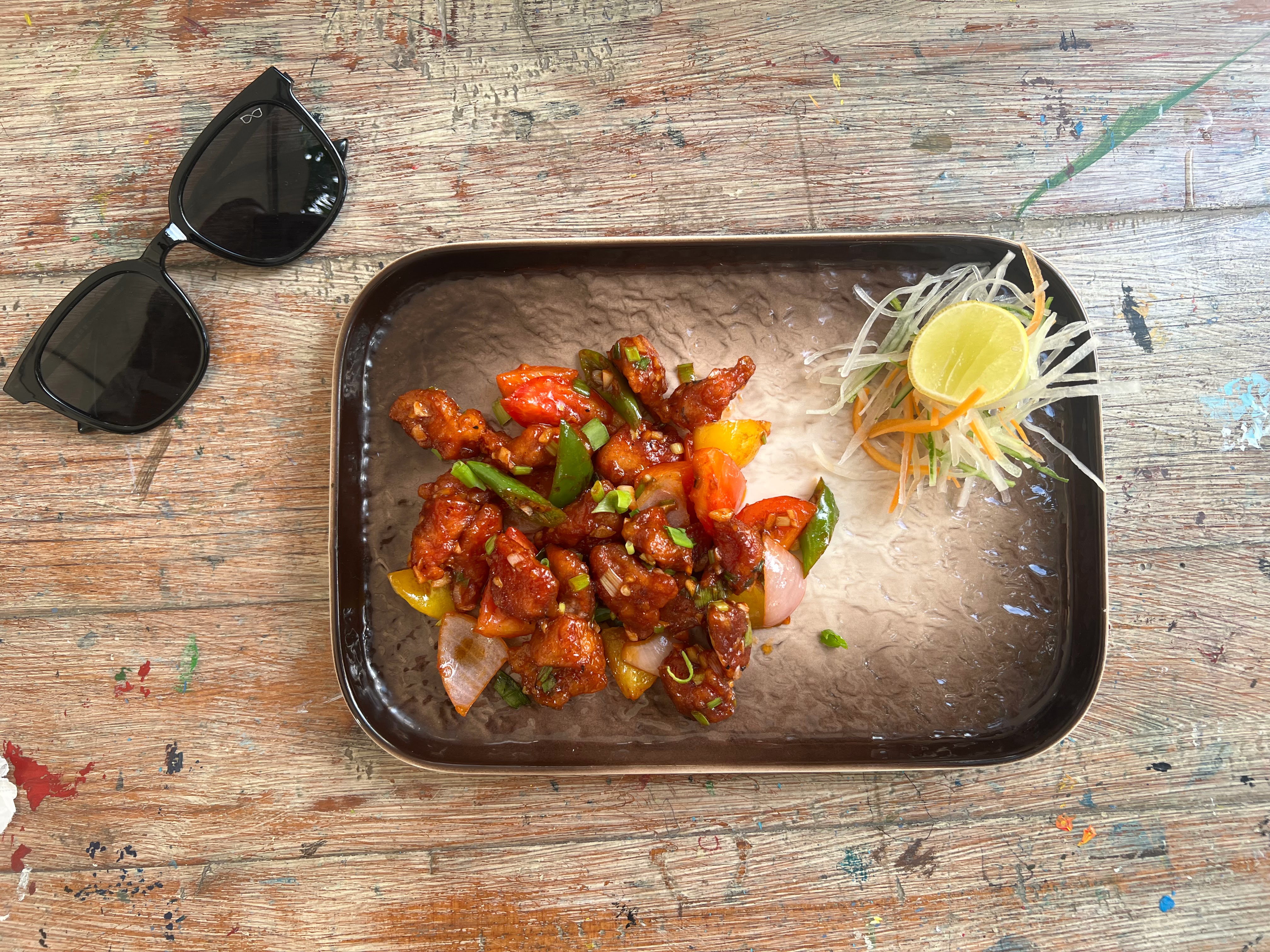 A rectangular plate holds spicy chili pork and bell peppers next to a small garnish of shredded vegetables and a lime wedge. A pair of sunglasses is on the left.