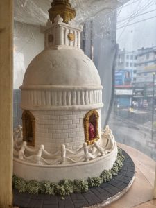 Highly decorative, very large mostly white iced cake, it resembles a circular dome topped statue or monument. A sculpted head, tile work and even a broken fence are included in the decorative detail.
